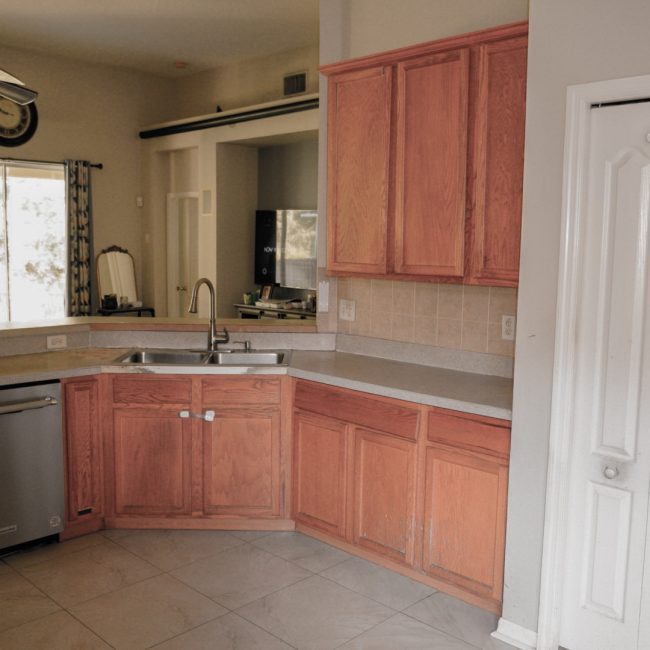 Before renovation of a kitchen with oak cabinets, white countertops, and a double sink.