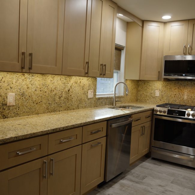 Completed kitchen renovation with tan shaker cabinets, granite backsplash, and stainless steel appliances.